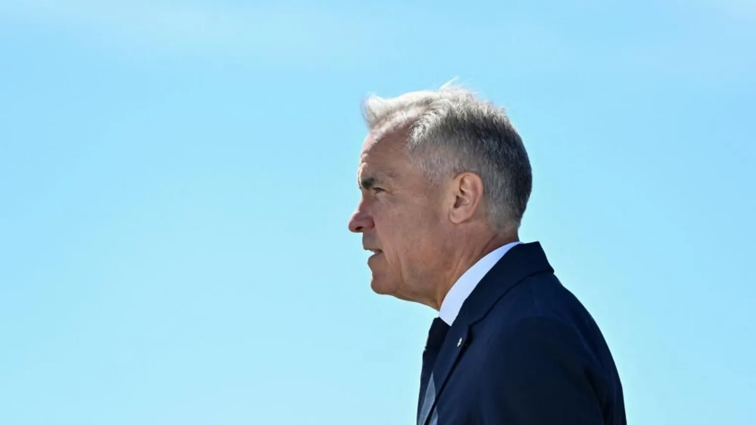 Reuters Mark Carney stands in profile, facing to the left, against a clear light blue sky. He wears a dark blue suit and tie.