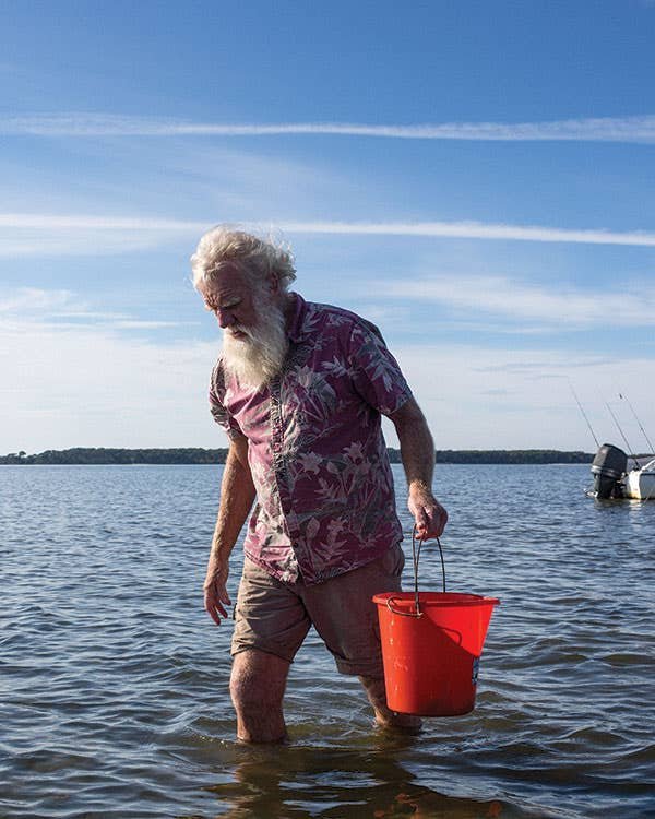 The Steward of Australia’s Original Food Older man with a white beard wades through shallow coastal water holding a red bucket, with a small motorboat anchored behind him.