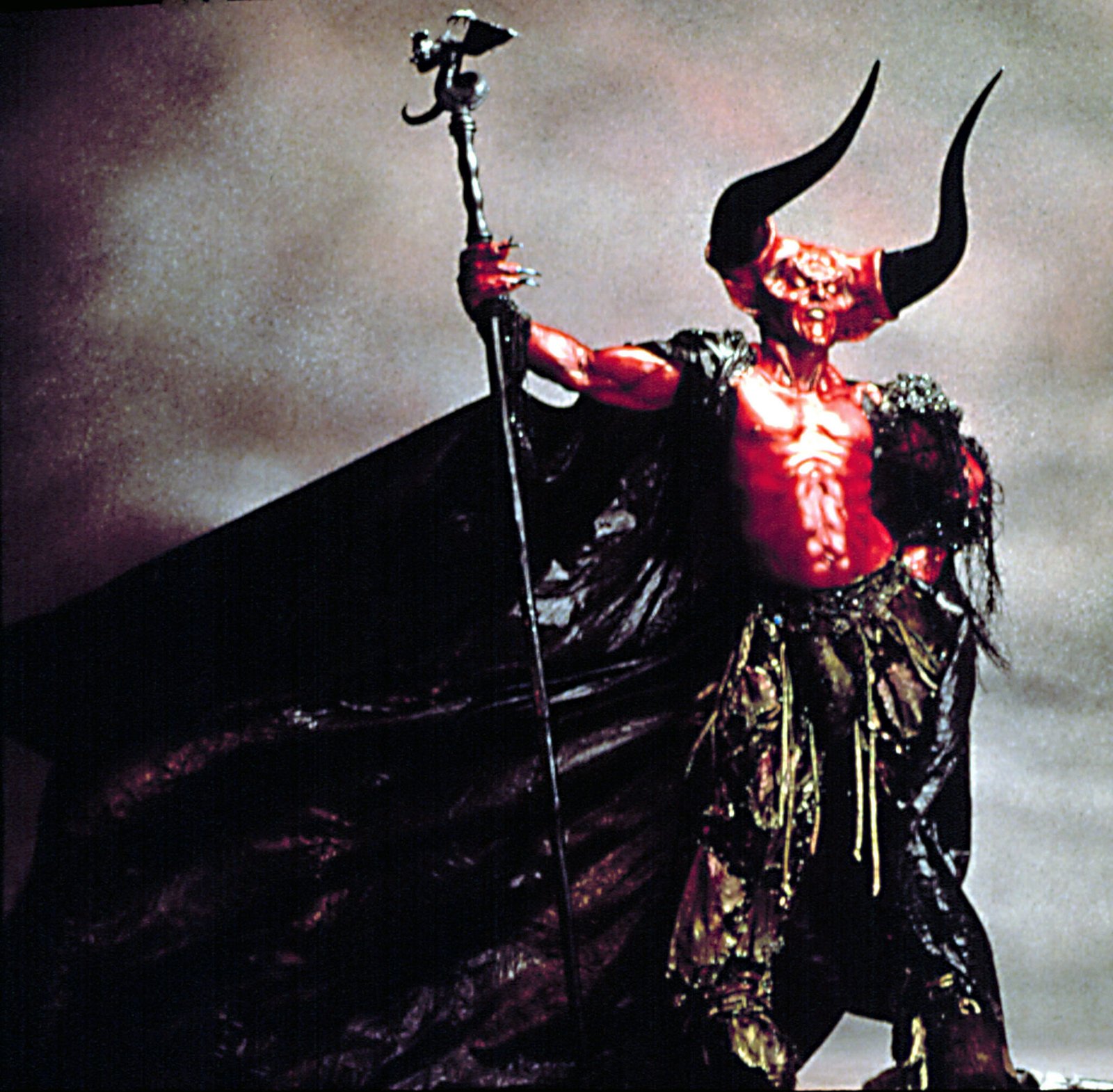 Tim Curry in full red, horned makeup and prosthetics as the Lord of Darkness in "Legend," holding a staff aloft, with a large black cape draped behind him.