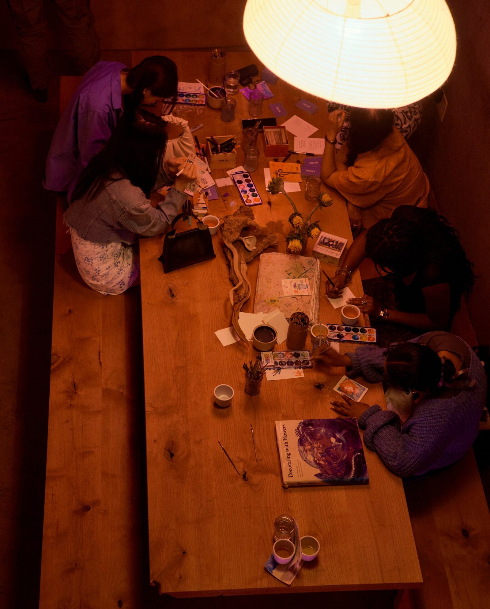 an overhead shot shows visitors at tea at shiloh painting with watercolors on a wooden table. a paper lantern hangs above them offering warm light.