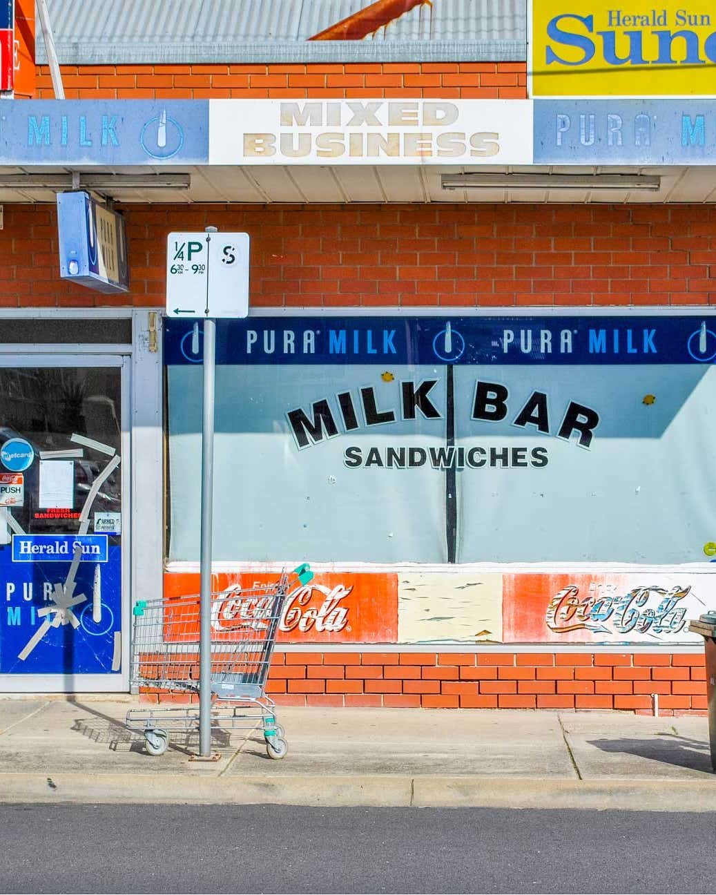 Remembering the Milk Bar, Australia’s Vanishing Neighborhood Staple Brick-front milk bar storefront with faded window lettering for milk bar sandwiches and pura milk, plus newspaper signage, a shopping cart on the sidewalk, and a trash bin nearby.