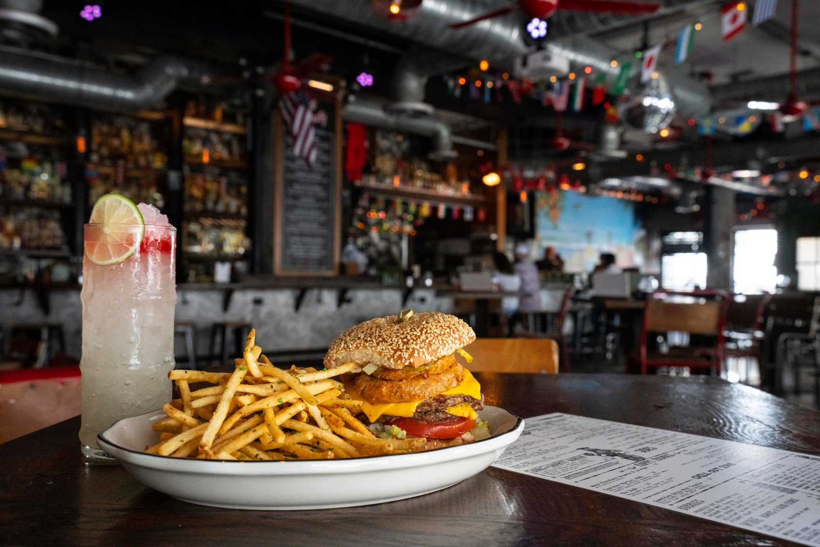 A hamburger and fries on a plate, a highball cocktail, and a menu on a table in a bar.