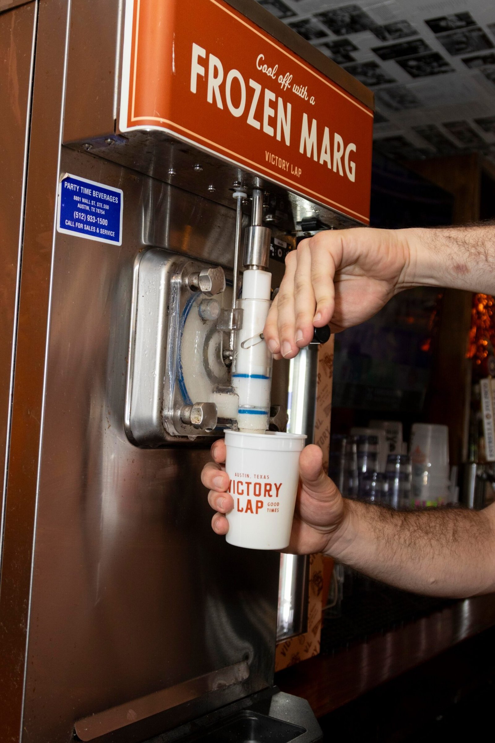 Hands operating a frozen margarita machine, pouring drink into a plastic cup with a logo that says Victory Lap