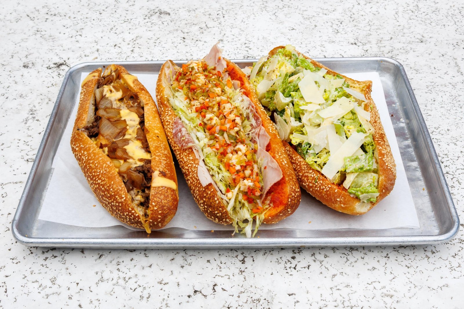 a lineup of three large hoagie-style sandwiches, a steak, an italian cold cut, and a chicken caesar on a silver tray