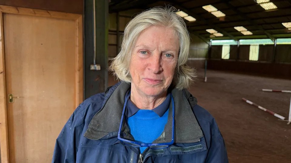 Lady with short hair that's light blonde. She is wearing a blue North Face coat. She has a blue polo shirt and blue jumper. She is standing in a stable.
