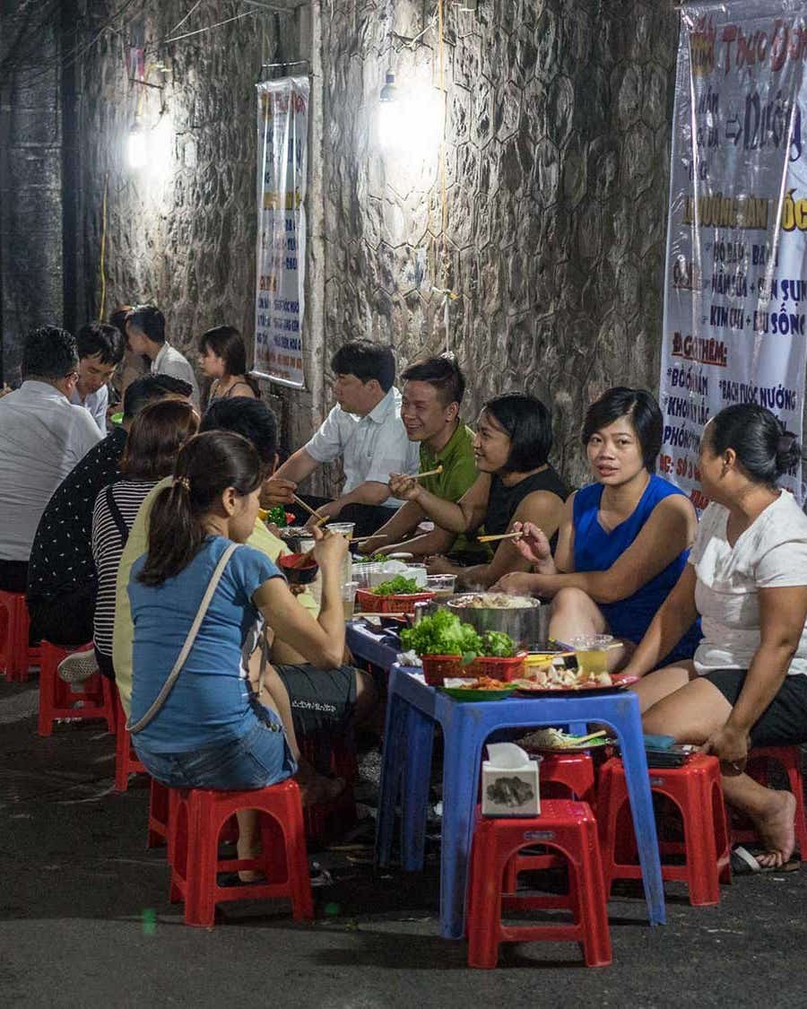 The SAVEUR Field Guide to Vietnamese Street Food Dishes Diners sit on small plastic stools at low tables along a narrow alley, eating bowls of Vietnamese street food under bright lights.