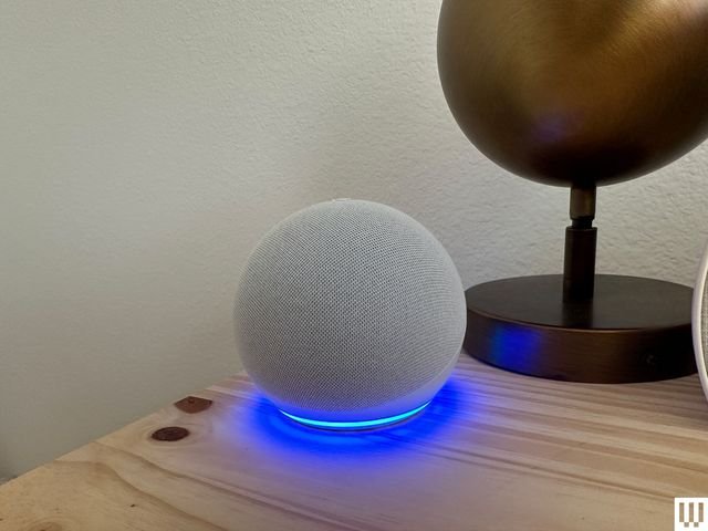 Front view of Echo Dot 5th Generation a grey sphere device with blue light illuminating from the base while sitting on a...
