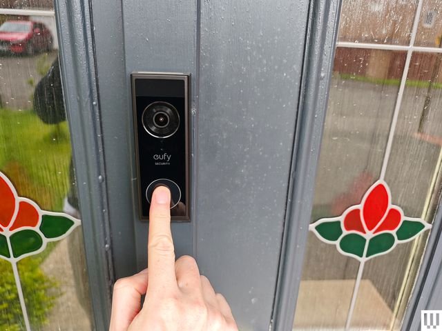Bronze rectangular electronic doorbell with builtin camera attached to a grey door while a person pushes the main button.
