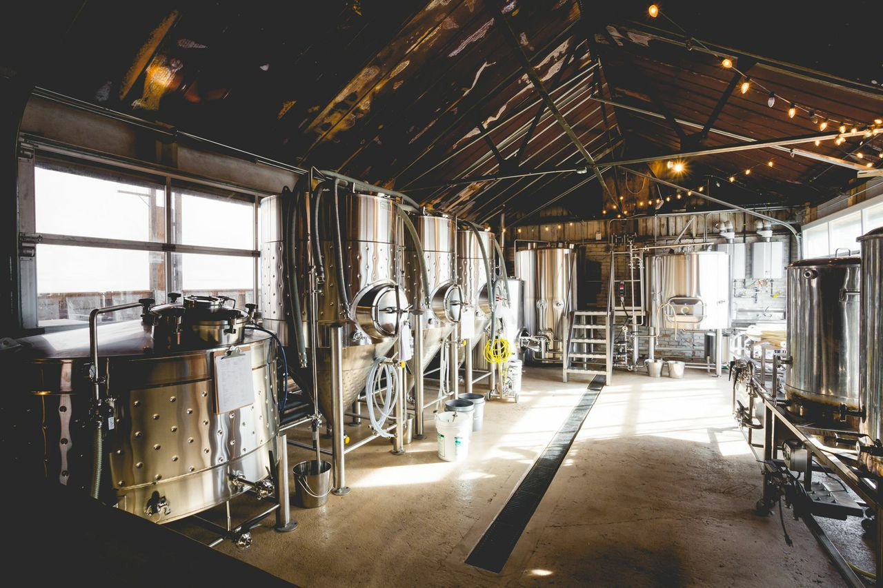 The 5 Breweries You Must Visit in Asheville, NC Stainless steel brewing tanks and equipment line the sunlit interior of a brewery beneath exposed rafters and string lights.
