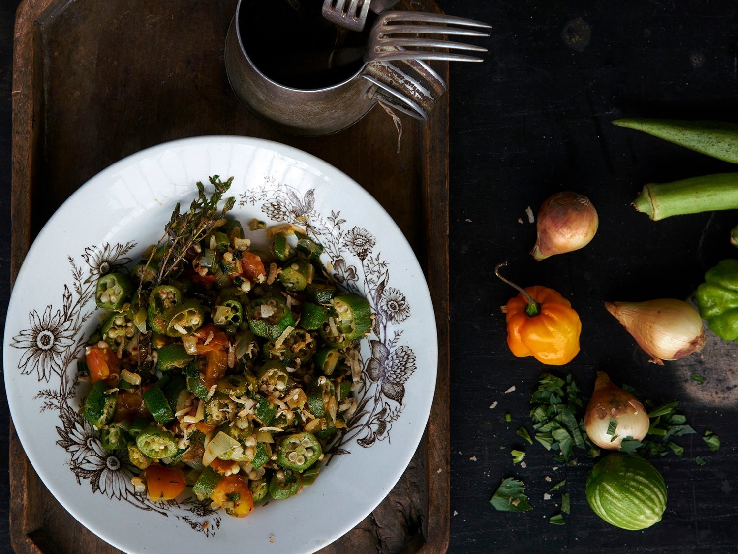 Sautéed sliced okra with tomatoes and onions, served in a patterned bowl on a wooden tray with forks; whole okra pods, shallots, a small orange chile, herbs, and a lime sit nearby.