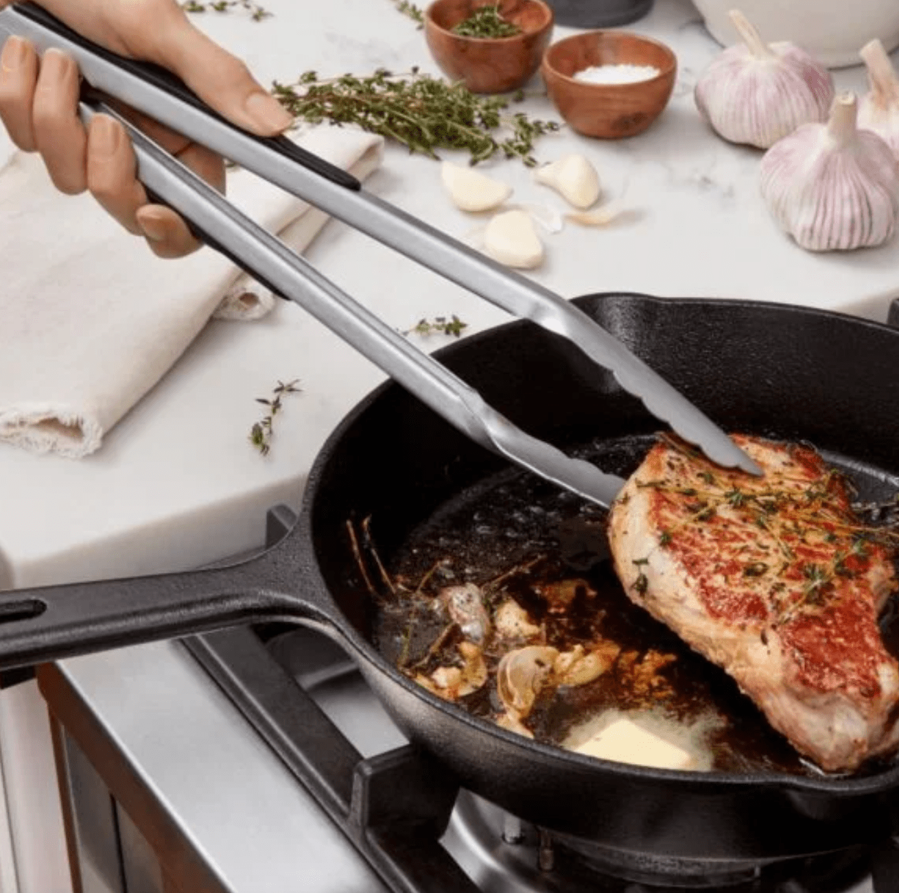 hand holding silver tongs gripping meat in a cast-iron skillet on a stove.