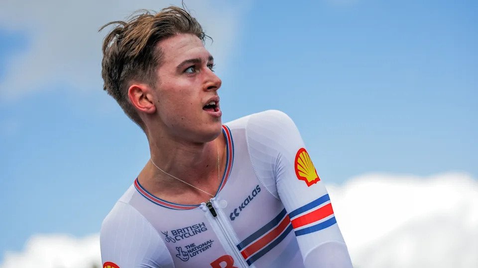 The side profile of a young man with brown hair and shorter sides looking to the side with his mouth partially open. He is wearing a white lycra top which says "British Cycling" on it. The sky is behind him.
