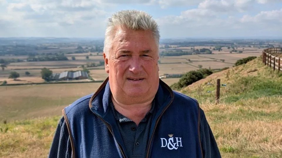 Paul Nicholls, a middle-aged man wearing a navy jumper with a collar is standing on a hill with grass fields in the background.