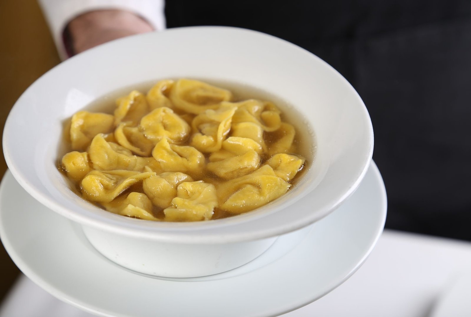 A server hands over a bowl of tortellini in broth on a saucer.