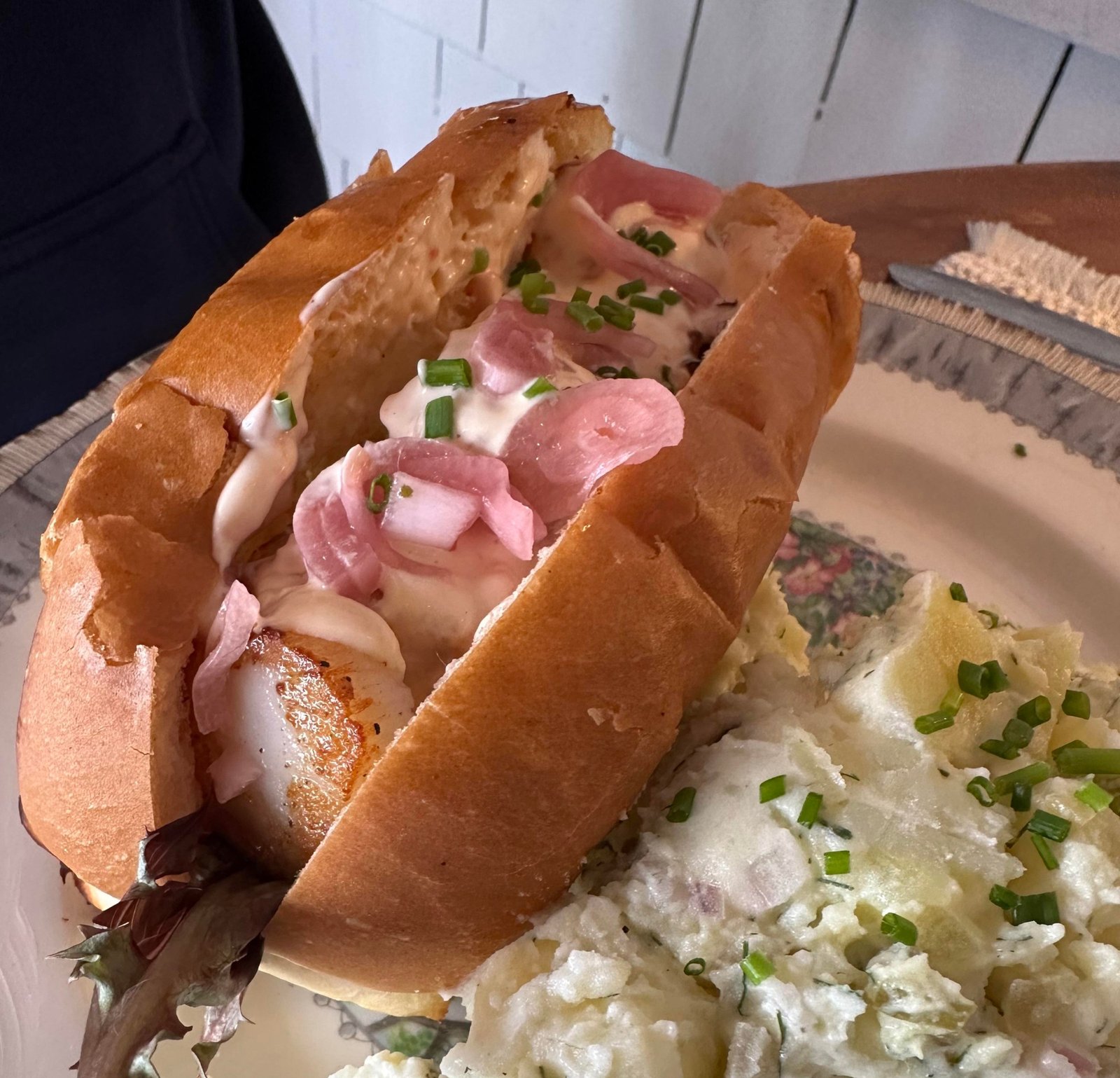 Scallops on a hot dog bun, garnished with pickled red onions and chives, with a side of potato salad.