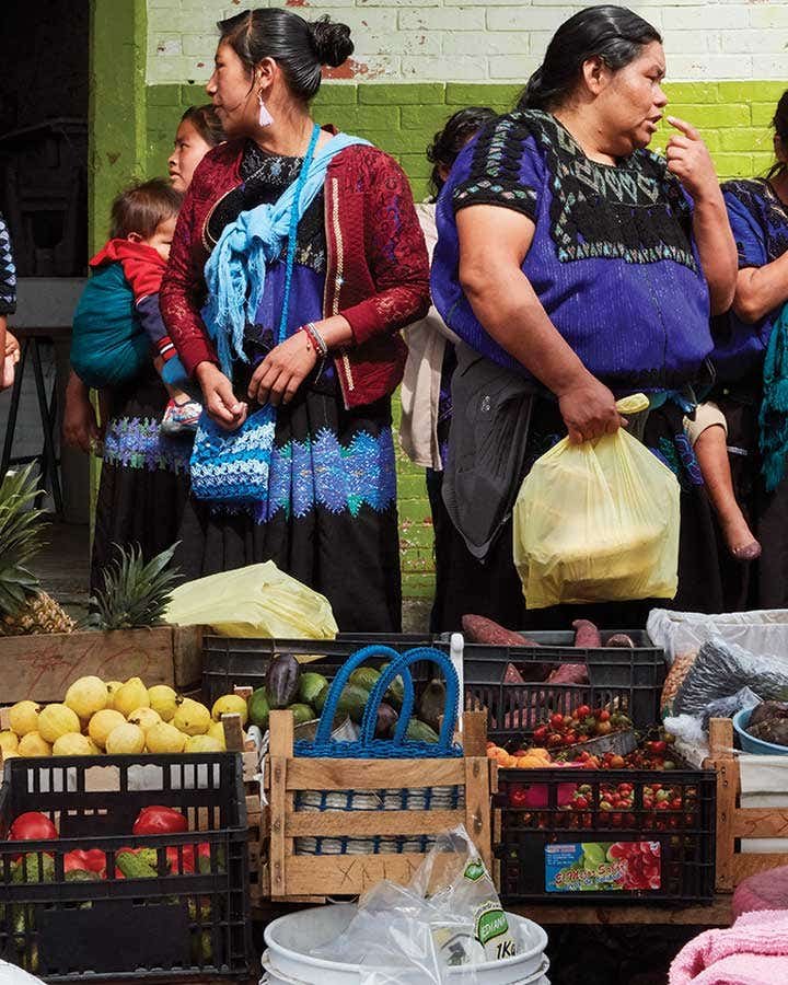 The “Women of the Corn” Share More than Maize at Yo’on Ixim Women and children in traditional woven clothing stand behind a market stall with crates of fruit and vegetables, including pineapples, tomatoes, and small red berries.
