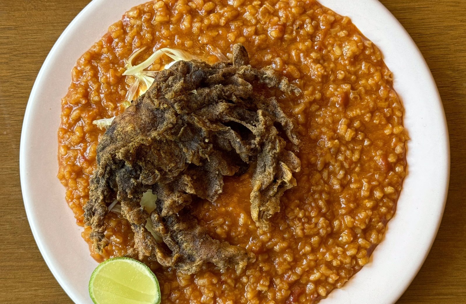 On a white plate on a wooden table, a deep-fried soft-shell crab sits atop saucy red rice with a wedge of lime next to it.