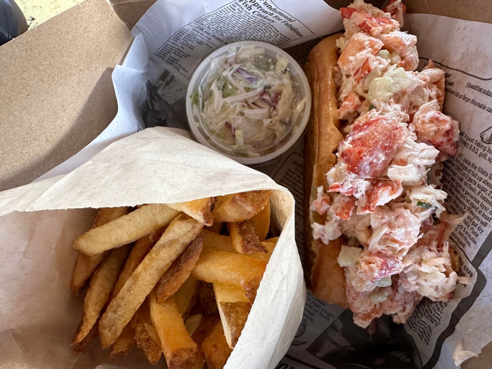 Lobster roll, thick cut fries, and a cup of coleslaw served on newspaper.