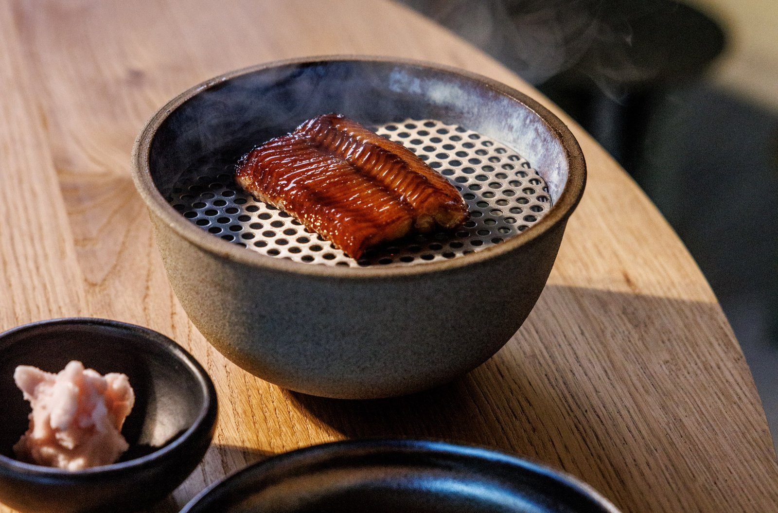 A lid lifts off a mini grill revealing glazed eel, presented beside pickled vegetables and another blurred dish.